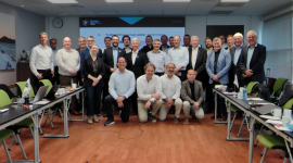 Group photo of about 30 professionals in a conference room, posing for a bannered workshop with a presentation screen in the back and laptops on tables on both sides.