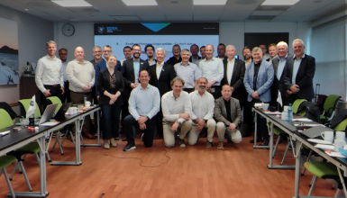 Group photo of about 30 professionals in a conference room, posing for a bannered workshop with a presentation screen in the back and laptops on tables on both sides.