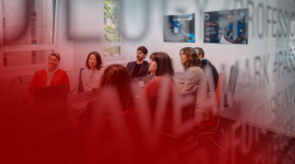 Group of professionals seated around a conference table in a glass-walled room, laptops open and screens displaying data as they discuss.