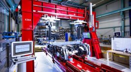 Red industrial machine in a factory with a computer control console at the left and metal workpieces on a transport bed. It appears to be a large gantry-type press or loader in a manufacturing area in operation.