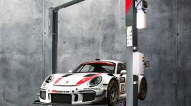 White Porsche race car with red stripe on a hydraulic lift in a concrete garage setting.
