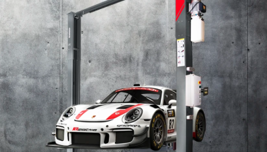 White Porsche race car with red stripe on a hydraulic lift in a concrete garage setting.