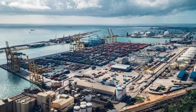 Busy container port with colorful shipping containers, large gantry cranes, and warehouses along the edge of the water