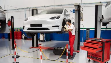 White car on a hydraulic lift being worked on by a mechanic in red overalls in a bright workshop