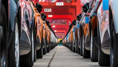 Rows of parked cars seen from ground level in a parking garage with red overhead beams and numbered signs above end of rows